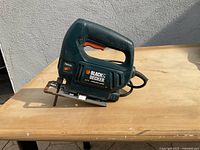 Green and black Black & Decker jig saw with orange trigger on wooden table against a white wall, showing the left side profile of the saw, cord visible