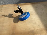 Photo shows blue circular clog cleaner with attached black handle and metallic hose connector on wooden surface with shadow cast.
