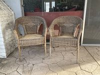 Photo showing two wicker chairs against a stone patio floor and glass door background. Chairs have a neutral, light beige and brown color tone.