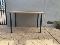 Full front view of the wooden table showing natural wood top and four black metal legs against a concrete patterned floor background