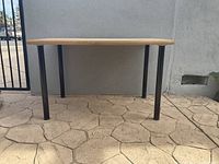Front view of the wooden table placed outside against a wall, showing the wooden top and black metal legs.