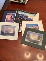 Photo showing five assorted artwork prints, some matted in different colors, depicting various scenes including cityscape, snowy village, coastal towns, and abstract art.
