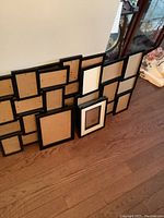 Multiple assorted black photo frames leaning against a wall on a wooden floor, showing backs of frames with backing and clips.