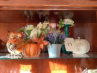 Shelf display featuring assorted artificial floral arrangements and fall-themed decorative pieces including ceramic pumpkin, owl figurine, white pumpkin with inscription, ceramic boot vase, and multiple artificial flowers in various containers.