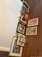 Nine assorted framed artworks arranged on wooden floor against white wall, showing various sizes and frame colors.