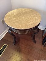One round end table with a beige marble top and dark curved legs on a wooden floor near wall vent