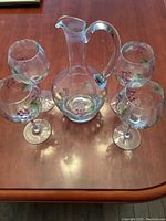 Full set of one glass decanter and four matching wine glasses with painted grape motifs arranged on wooden table.