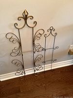Two wrought iron decorative hanging hardware units positioned against a wall on hardwood floor, showing detailed scrollwork design and shape differences.