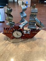 Photo of a decorative ship themed clock with metal sails and wooden hull base, showing the clock face and compass on the hull, placed on a beige surface.