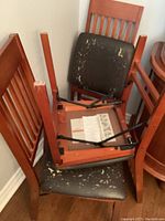 Three wooden folding dining chairs stacked against a wall showing peeling and damaged black vinyl seat cushions and slatted backs.