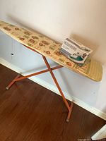 Ironing board with patterned cover and orange legs, with boxed iron on top