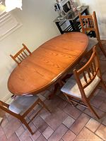 Photo showing wooden oval kitchen table with four matching chairs on tiled floor, table leaf installed making oval shape.