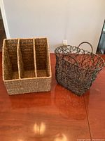 Two magazine racks placed on a wooden table against a white wall. One is a woven natural fiber rack with three compartments, the other a metal wire basket with handles and an open mesh design.