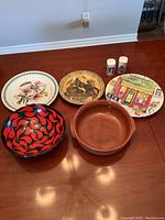 Photo of five decorative pieces on wooden surface including two painted plates, two bowls, and a pair of salt and pepper shakers