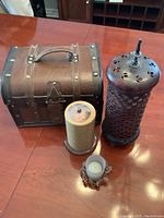 Photo showing four decorative items on a wooden table: a small wooden chest, a tall ornate metal container with lid, a beige candle on a base, and a small metal candle holder with a candle inside.