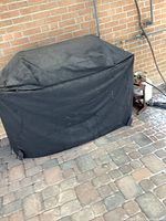 Grill covered with black protective cover against brick wall, propane hose and regulator stand visible to right