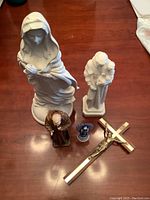 Photo with six religious statues and one brass crucifix on wooden table showcasing size and material variety.