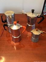 Four varying sizes of aluminum stovetop espresso makers (Moka pots) on a wooden table, showing black and orange handles and knobs.