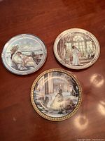 Three decorative plates featuring vintage romantic scenes by Trisha Romance, placed on a wooden surface.