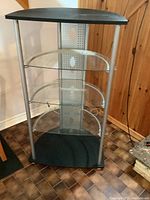 TV audio video stand from front showing black top and base, three glass shelves, and silver metal back panel with cable holes.