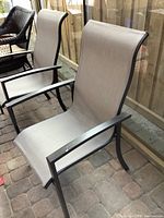 Two beige mesh and metal frame patio chairs placed on a brick surface near a glass window and other outdoor furniture.