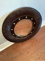 Photo of a round wall mirror with a dark brown textured frame and brass rivet accents, showing the mirror leaning against a wall on a wooden floor.