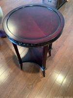 Wood round side table with carved detailing on the edge, four legs and a lower shelf, shown on wood floor.