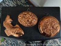 Three copper molds arranged on a black surface: fish shape, oval, and round with embossed designs and hanging loops