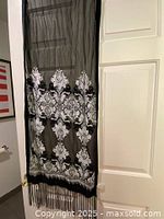 Black and white sheer scarf with stitched floral and ornamental patterns, hanging on door, showing fringe details