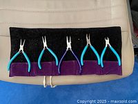 Five jewelry making pliers with blue handles laid out inside purple fabric roll-up case, handles facing out, tips facing in.