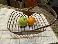 Rectangular metal wire basket with wide spaced metal rods and metal handle on a checkered kitchen towel, holding apple and orange for scale.
