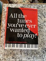 Front cover of 'All the Tunes You've Ever Wanted to Play' piano book with red and black cover and piano keyboard graphic