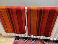 Two mid-century drapery panels displayed over railing, showing vibrant horizontal stripes in red, orange, tangerine, and brown, folded at the hem.