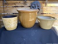 Three ceramic garden pots in varying cream and sand shades displayed on a dark surface with a decorative wall background.