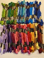 Photo shows a large assortment of skeins of DMC embroidery yarn arranged in rows grouped by color including greens, blues, purples, pinks, reds, oranges, yellows, and dark colors.