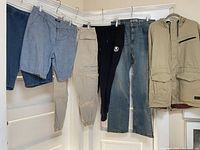 Six pieces of menswear hanging on wall corner including pants, sweatpants, jeans and jacket.