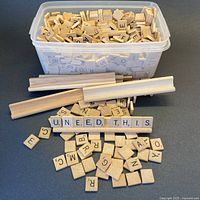 Plastic container filled with hundreds of wooden Scrabble letter tiles with four wooden racks and scattered tiles in front, spelling 'UNEED THIS'.