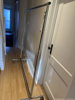 Full view of the metal rolling clothing rack showing the horizontal bar, vertical supports, and caster wheels in a narrow corridor.