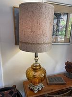 Full view of the mid-century modern lamp on a wooden surface, showing the amber blown glass body, ornate silver-tone metal base, and original tan-colored drum shade with cinnamon speckled accents.