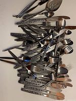 Photo showing a pile of various pieces of cutlery and flatware with textured embossed floral handles. Pieces include spoons, forks, and knives of various sizes, arranged overlapping.