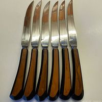 Full set of 6 serrated cutting knives with black and cinnamon Bakelite handles arranged in parallel to show blade edges and handle detail.
