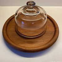 Angled side view of round solid teak cheese board with fitted clear glass dome cover set on top, showing natural wood grain and dome knob.
