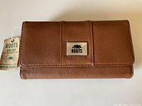 Front view of the closed caramel leather ROOTS wallet with metal logo and attached RFID safe tag.