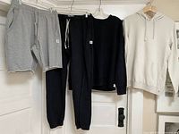 Navy blue sweatpants with white drawstrings, matching navy sweatshirt, cream hoodie, and grey sweatshorts displayed hanging on hangers.