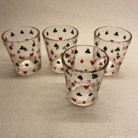 Front view of 4 clear glasses decorated with red and black playing card suits (clubs, spades, hearts, diamonds) evenly spaced around each glass