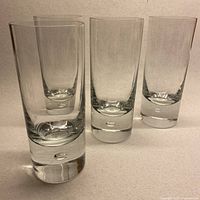 Four clear glass highball tumblers with thick bottoms and visible bubbles embedded in the center of the glass base.