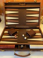 Open backgammon board showing felt playing surface with cinnamon and cream points, dice cups, dice, playing pieces, doubling cubes, and wooden carrying case with leather handle.