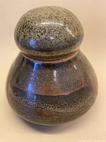 Front view of vintage studio pottery lidded vessel showing rounded shape, muted earth-tone speckled glaze with dark horizontal bands.