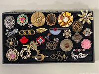 Full top view of the box with over 20 assorted vintage costume brooches in various designs and colors.