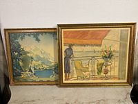 Two framed prints shown side by side against a wall on a white cloth surface, one interior scene, one scenic lake and mountains.
